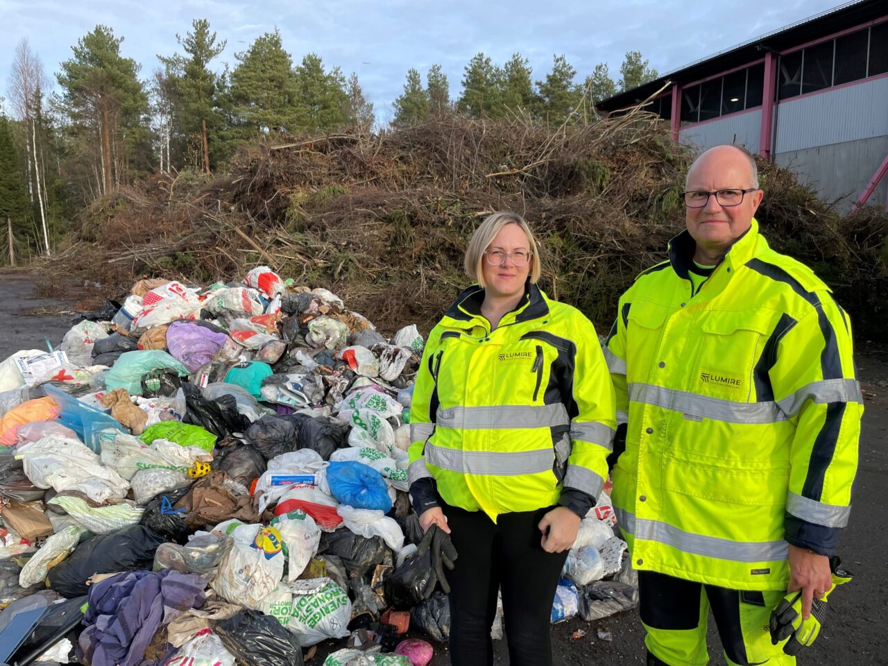 Två människor i gula arbets- och varselkläder utomhus vid en stor soptipp med olika påsar och avfall