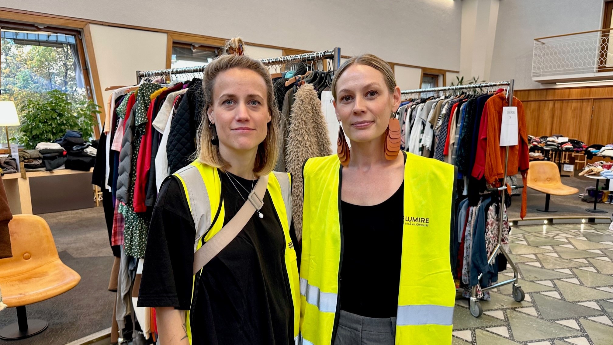 Två människor i gula varselvästar inomhus vid klädställningar