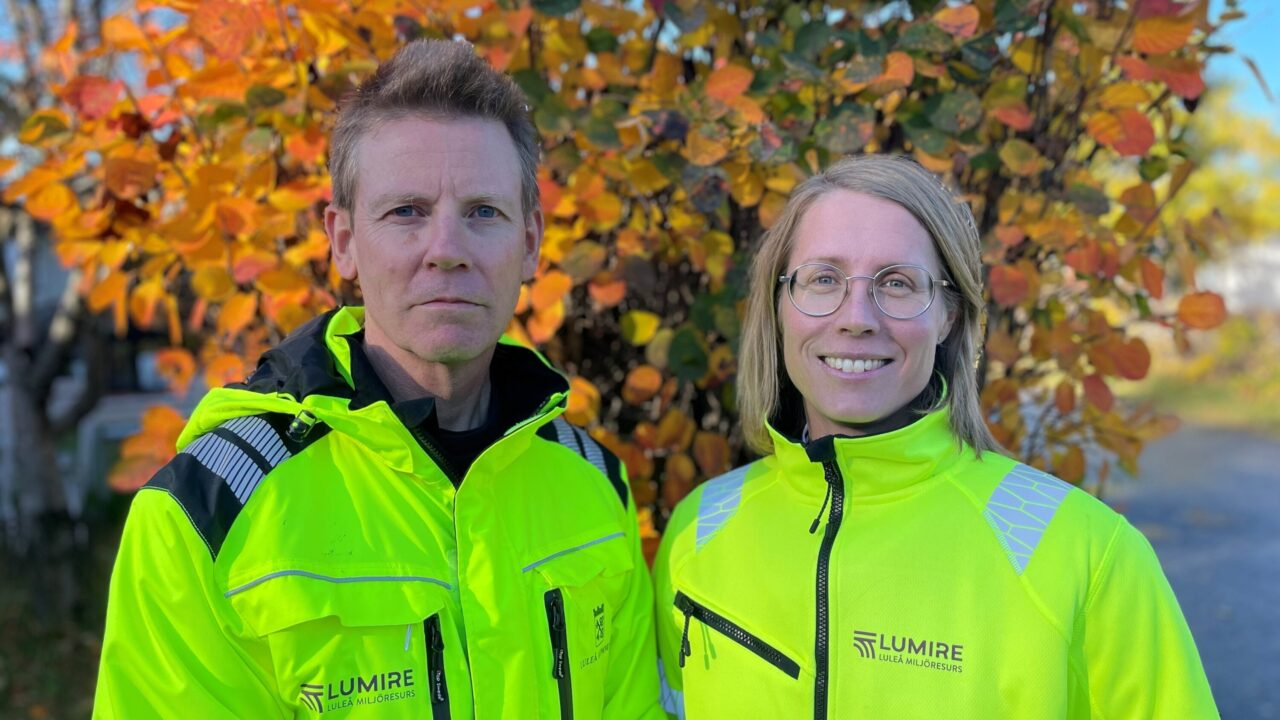 Två människor i gula arbetskläder utomhus vid höstfärgat träd