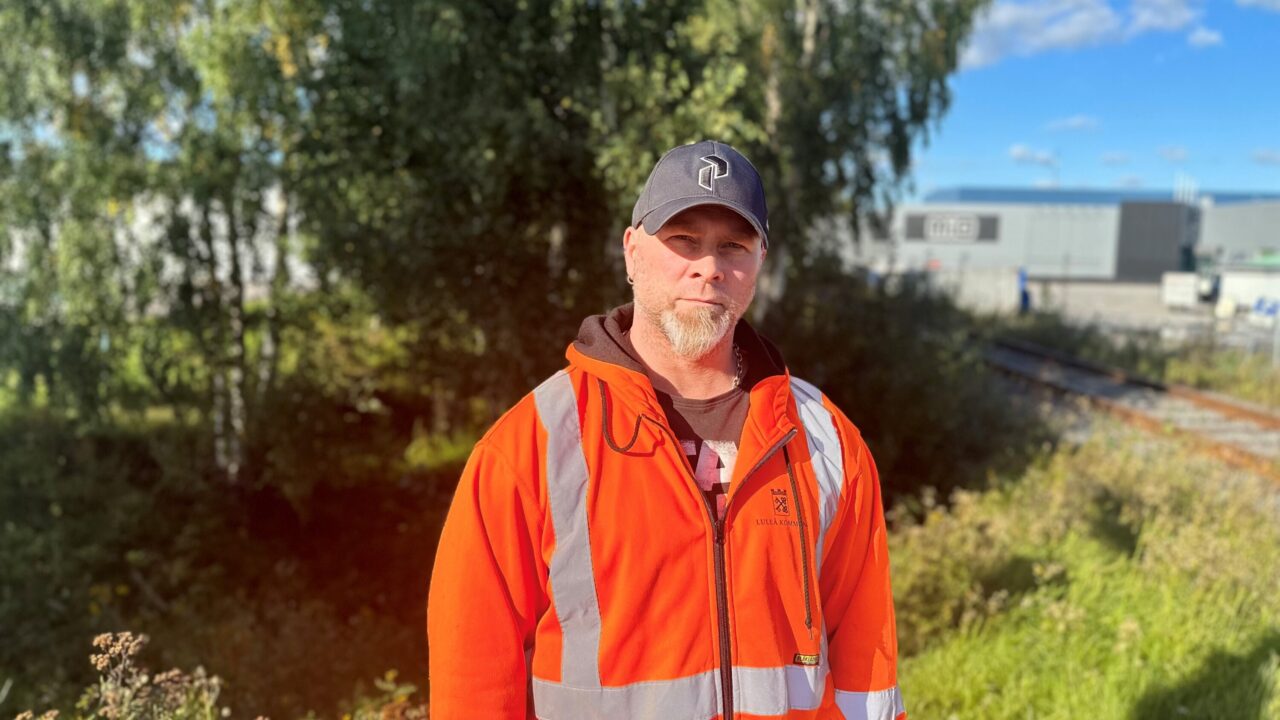 Bild på människa i orange arbetskläder vid grönskande plats med järnväg i bakgrunden