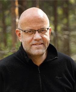 Bild på en person som står framför en skog. Personen tittar rakt in i kameran.