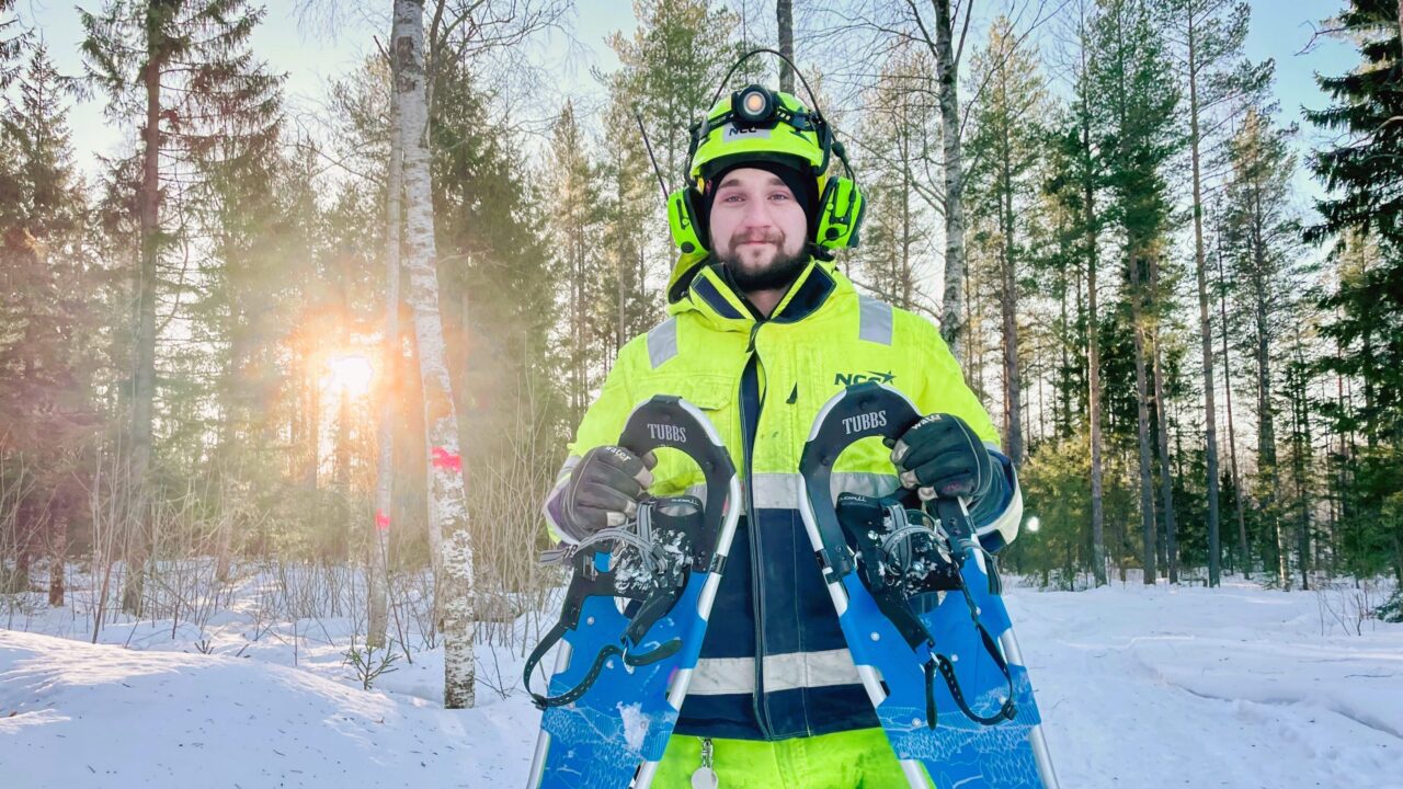 Människa utomhus i snö och sol med snöskor i handen, och med gula arbetskläder på sig