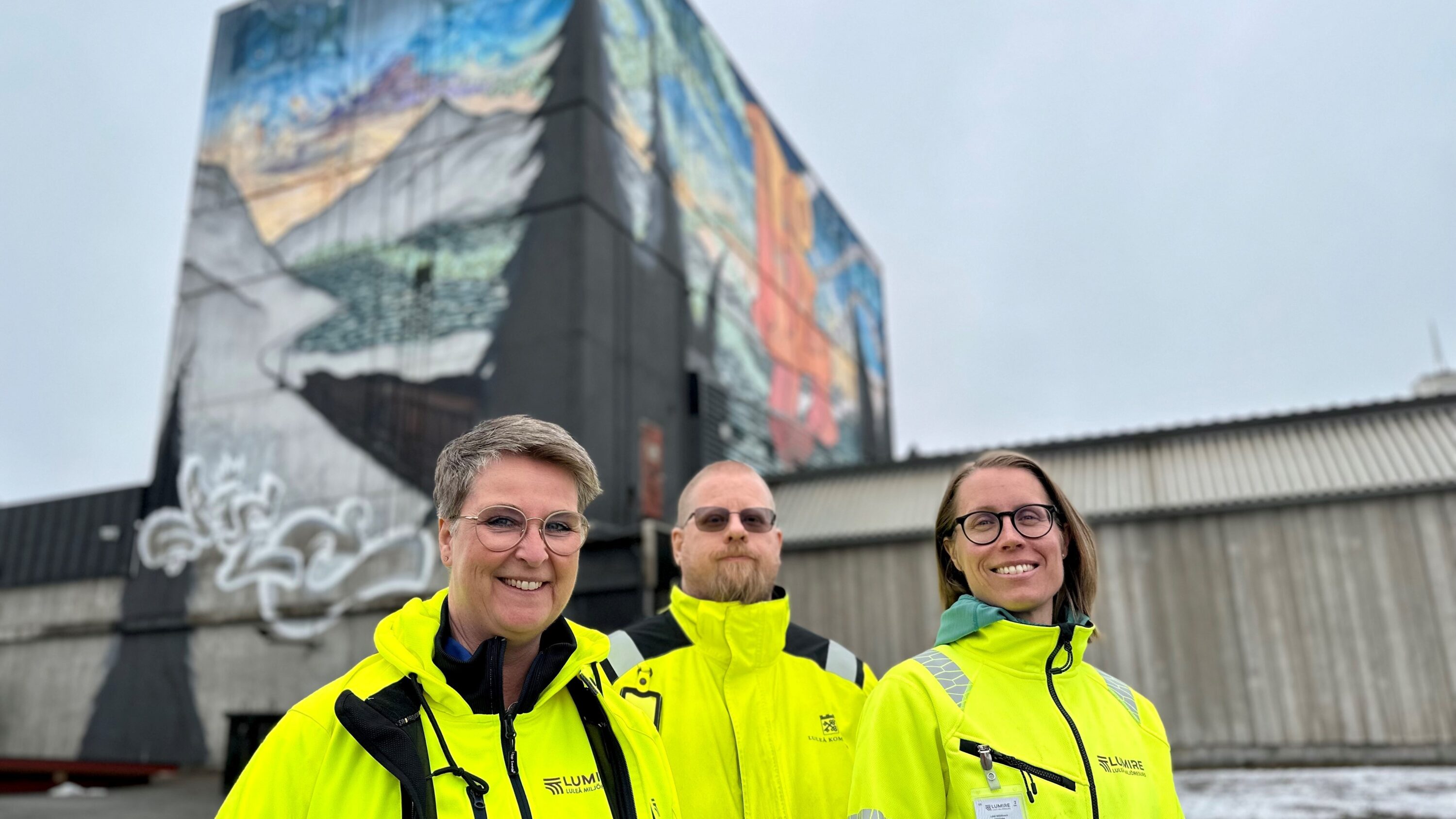 Bild på tre personer utomhus i gula arbetskläder framför industribyggnad med grafittimålning