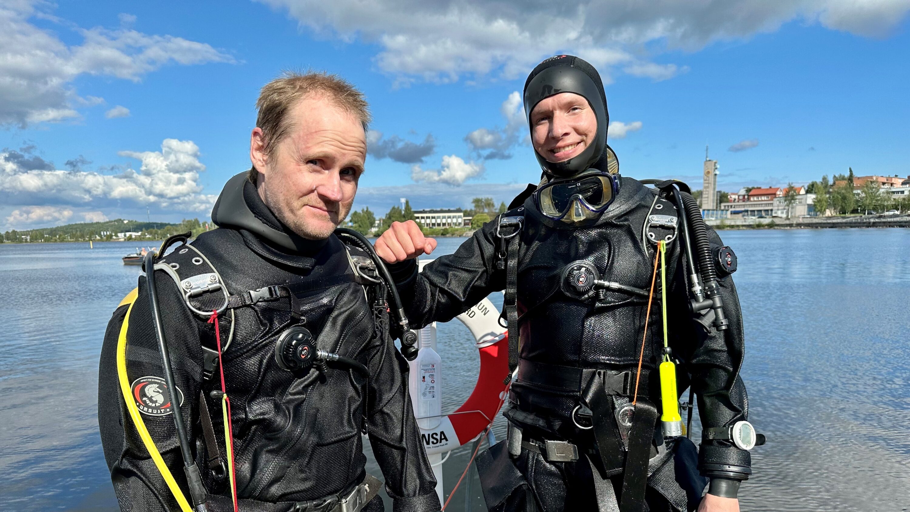 två dykare just uppstigna i dykarutrustning på en brygga vid solig dag blå himmel vita moln