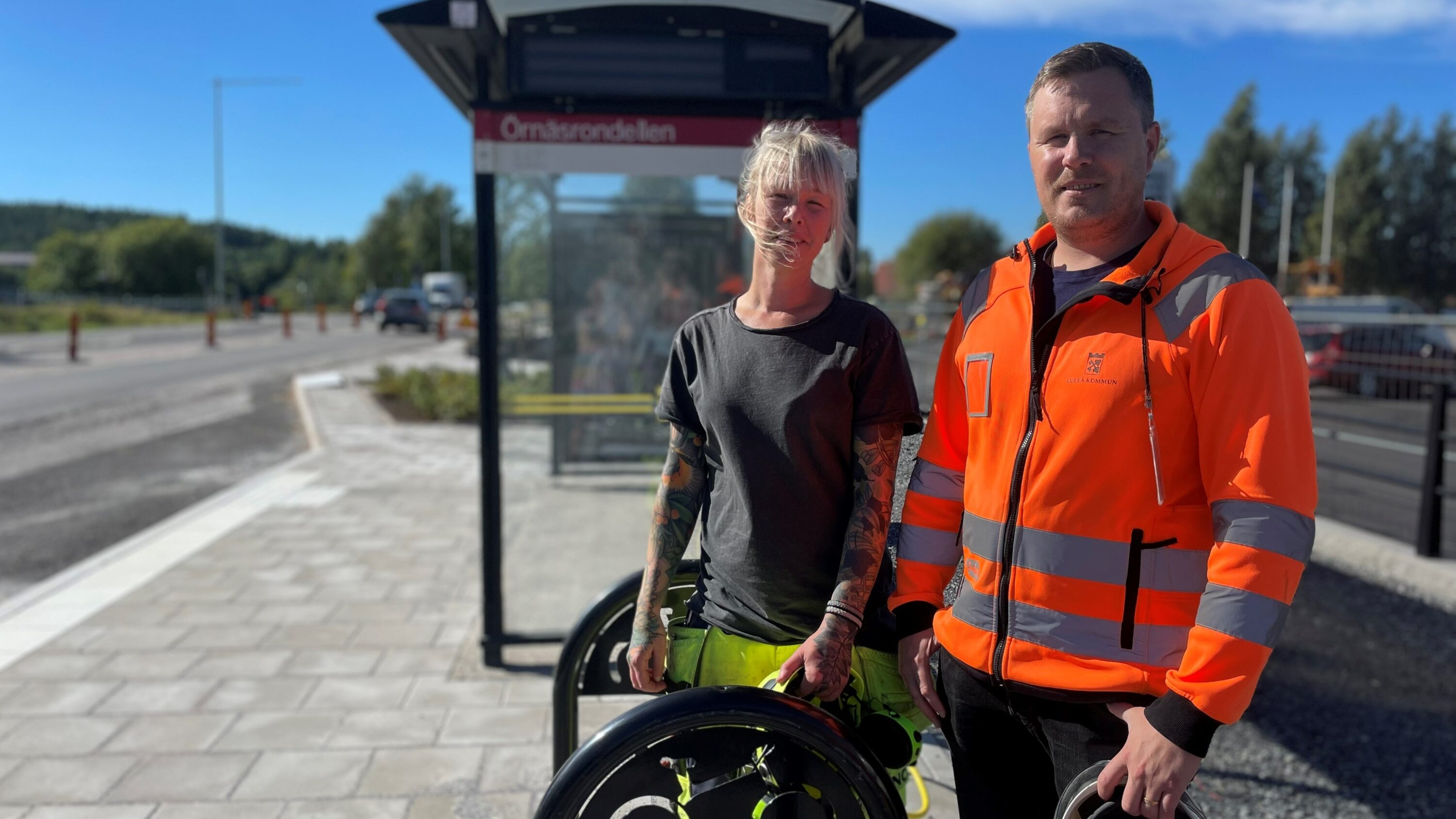 Två människor utomhus i sol i skyddskläder vid busshållplats