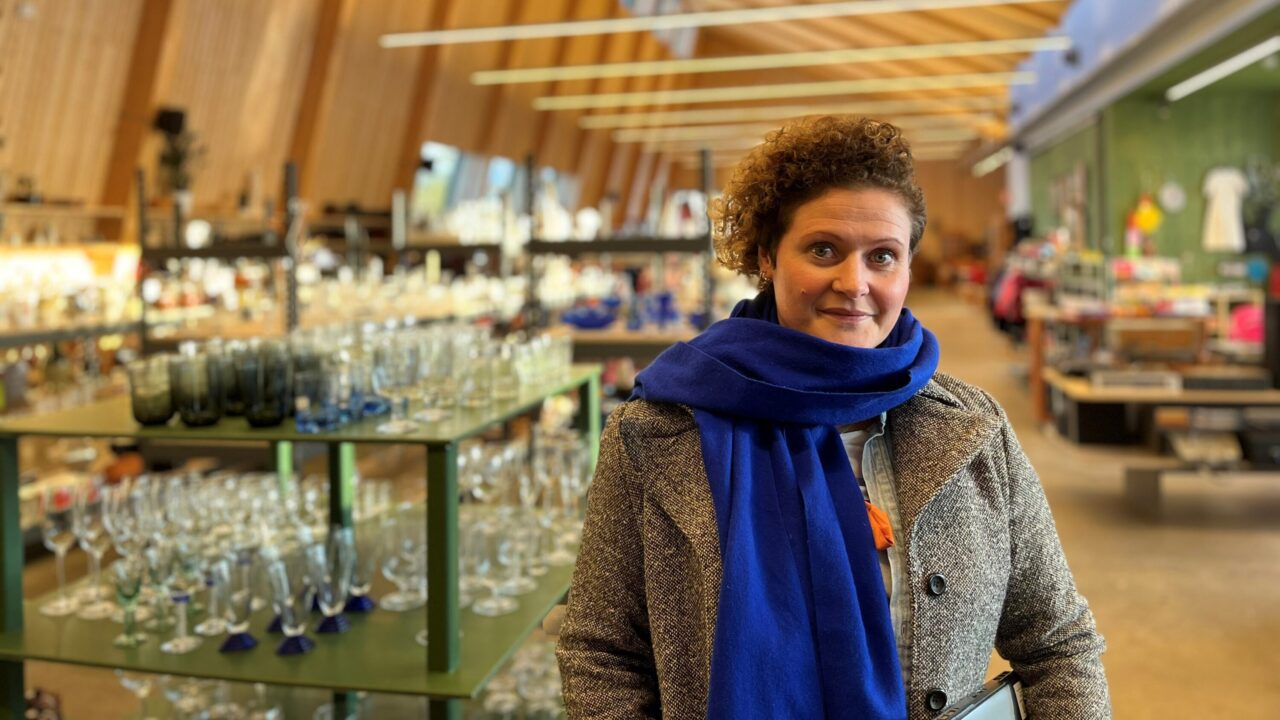 Bild på människa i vinterklädermed blå halsduk inomhus i trähus med glasföremål på hyllor
