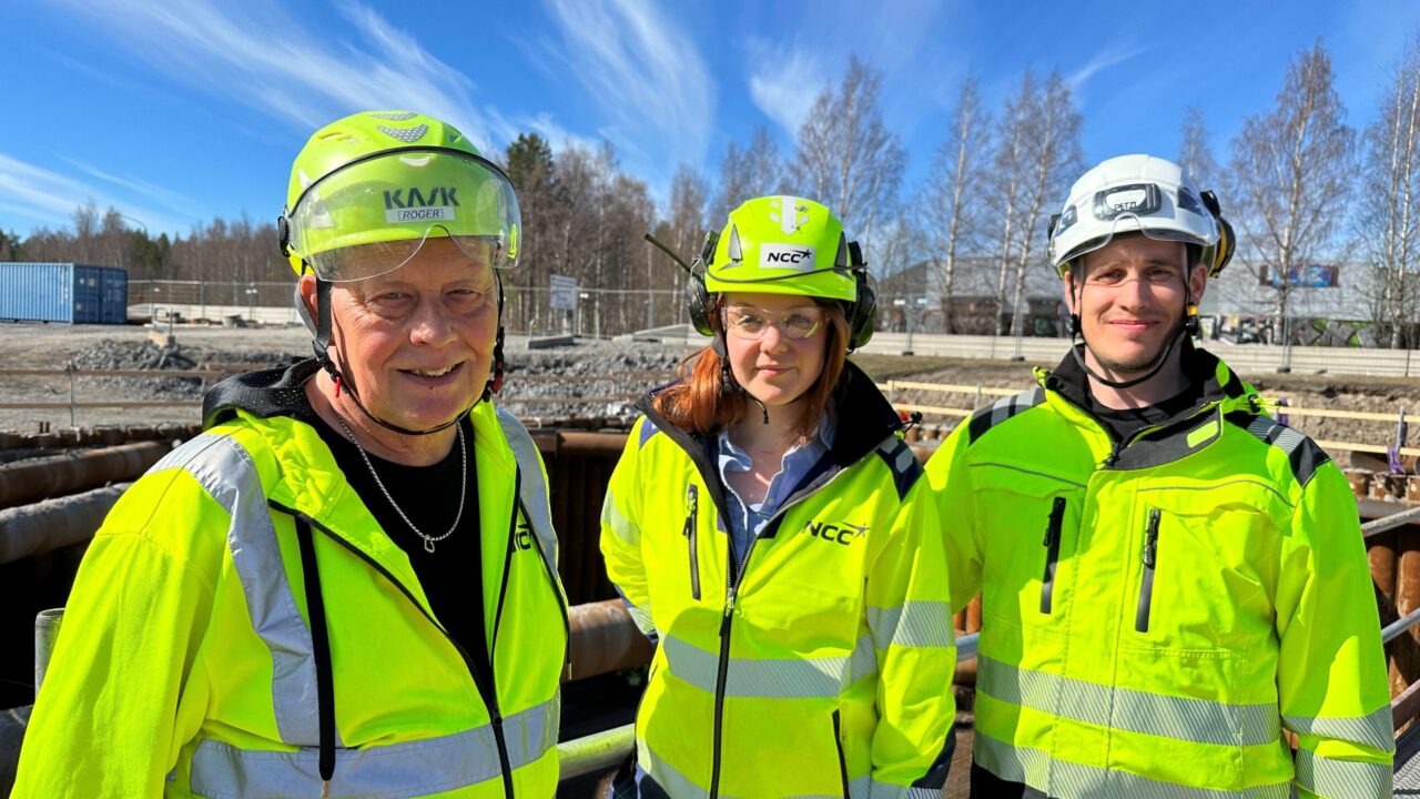 Tre personer i gula skyddskläder och hjälmar vid ett bygge i sol med blå himmel