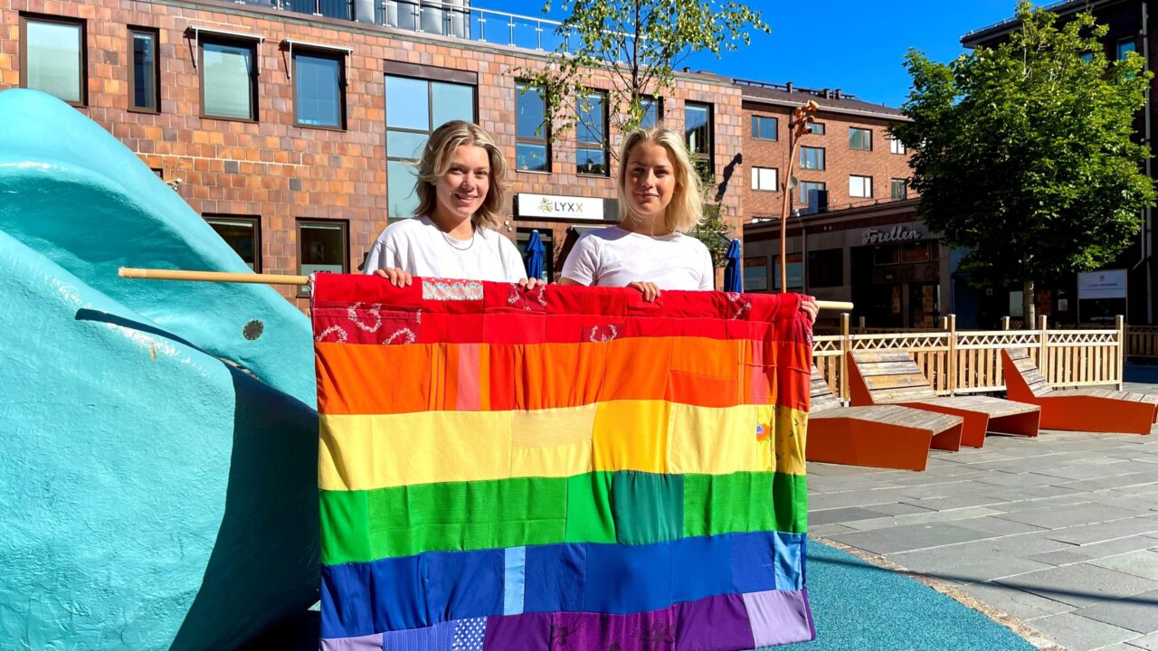 två människor håller i stor regnbågsflagga utomhus på ett torg