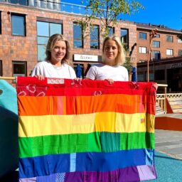 två människor håller i stor regnbågsflagga utomhus på ett torg